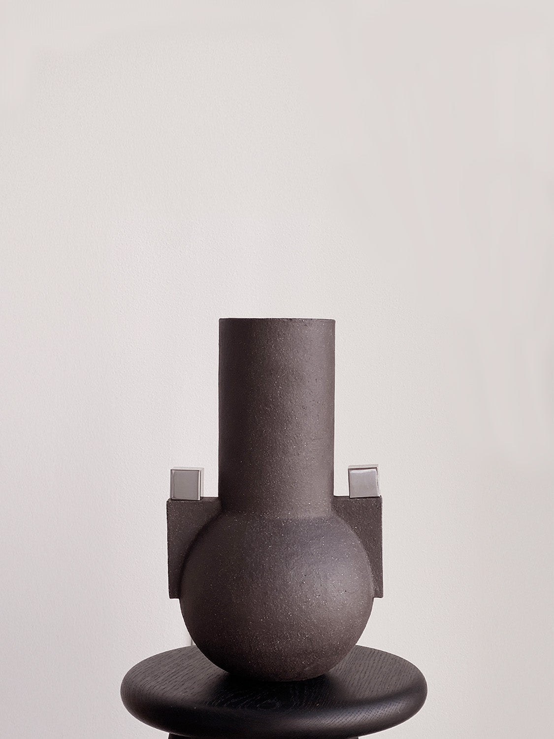 Dark gray vase with a cylindrical top and round base on a matching stand against a light gray background