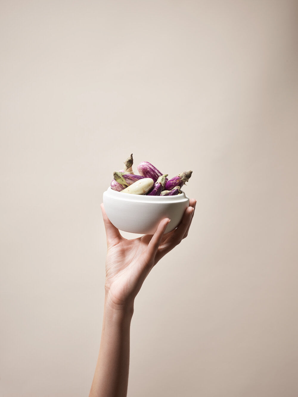 Hand holds up white matte earthenware container filled with baby eggplants